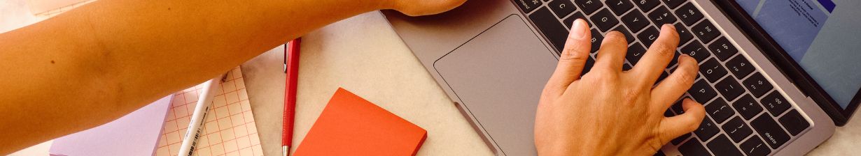 Hands typing on a laptop beside a notebook and pen on a desk